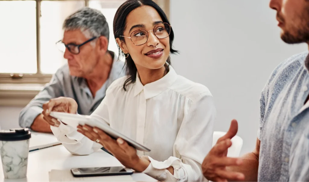 Photo of a woman speaking with other professionals in an office while holding a tablet.