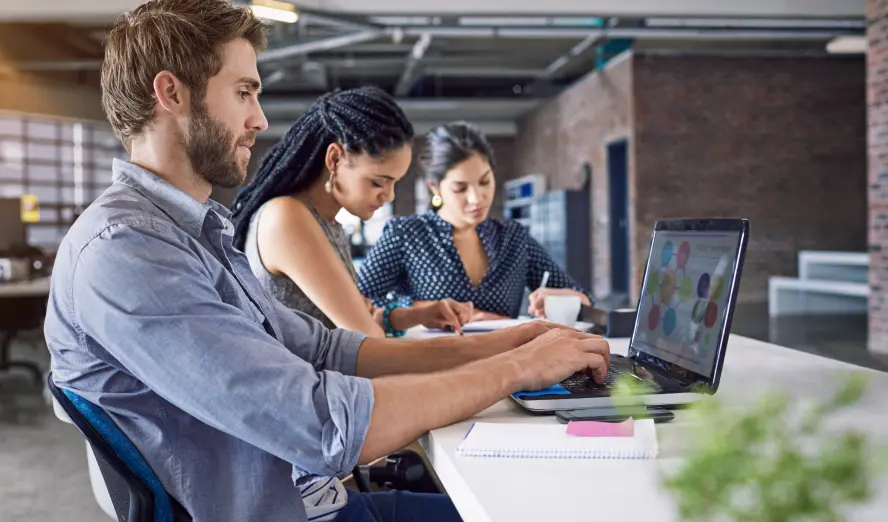 Photo of three people collaborating on a laptop in a professional office setting. Photo of three people collaborating on a laptop in a professional office setting.